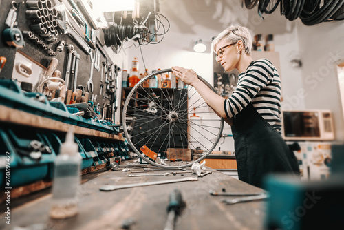 Fotomural Cute Caucasian female worker holding and repairing bicycle wheel while standing in bicycle workshop