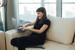 © makarovada - Happy smiling hipster girl typing on a laptop computer keyboard, chatting with family or  friends via online messenger while sitting on a cozy sofa ot home with modern interior design