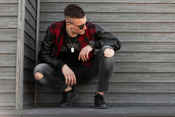 Sticker - Pretty handsome young hipster man in black sunglasses in trendy red checkered jacket in sneakers in stylish ripped jeans is sitting outdoors near a wooden wall. American guy is resting.