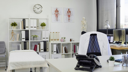 Doctor office table and chair with white lab coat on it and white paper nobody. telephone and tools on work desk by the therapy bed in clinic. bright and modern empty workplace in hospital.