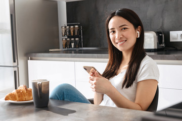 Sticker - Portrait of adorable asian girl holding mobile phone while having breakfast in kitchen at home