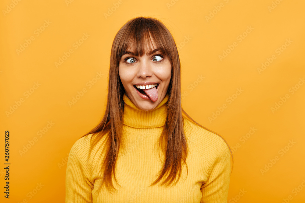 Woman portrait. Fun. Cheerful young woman is grimacing and showing her tongue at camera, on a yellow background - obrazy, fototapety, plakaty 