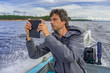 © Tatyana_Andreyeva - Handsome middle-aged man sitting at boat stern, floating along northern lake and using smartphone on beautiful landscape background in summer day. Travel concept. Onega lake, Karelia, Russia
