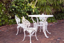 White Wrought Iron Table And Chair Set On Brick Patio In Tropical Garden