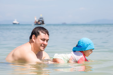 Father holds smiled and happy child in the water and teaches to swim. Summer vacation on tropical beach. Infant baby girl 9 month first time in the sea