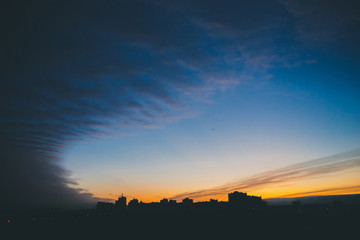 Cityscape with wonderful varicolored vivid dawn. Amazing dramatic blue cloud sky above dark silhouettes of city building roofs. Atmospheric background of orange sunrise in overcast weather. Copy space