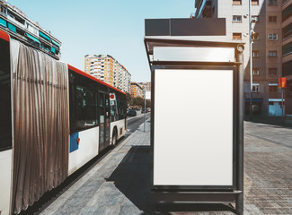 An empty ad poster mock-up on an outer side of the bus stop; advertising billboard placeholder template on a stop of public transport with a bus on the left; mockup of a blank white outdoor banner