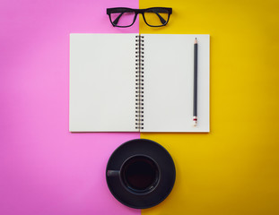 Blank white notepad with black coffee cup, pencil and glasses on two tone pink and yellow background.