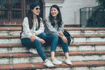 Wall Mural - Two cheerful asian girls talking and sitting on stairs in street Santa Barbara County Courthouse. concept of sincere friendship travel together lifestyle. female college students laughing joyful.