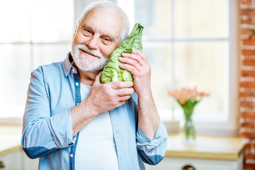 Wall Mural - Portrait of a cheerful senior man with fresh local grown lettuce at home on the window background