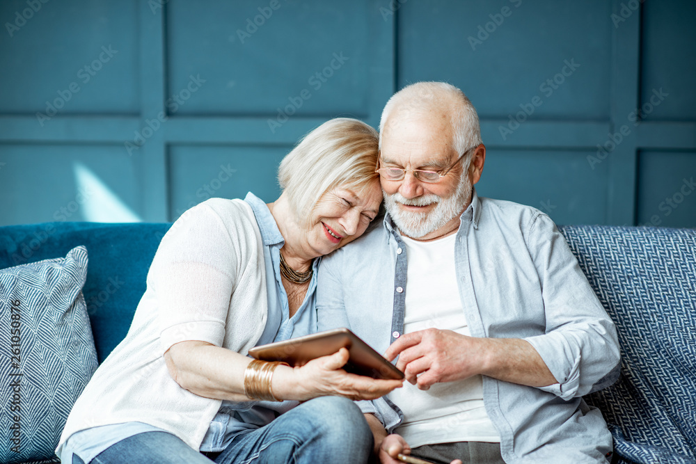 Lovely senior couple dressed casually using digital tablet while sitting together on the comfortable couch at home - obrazy, fototapety, plakaty 