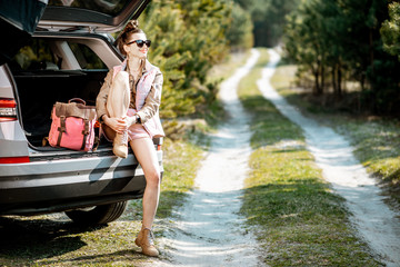 Wall Mural - Young woman enjoying nature while sitting in the car trunk on a picturesque road in the woods