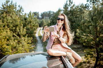 Wall Mural - Young woman enjoying a trip, photographing with phone while sitting on the car roof on a picturesque road in the woods