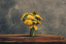 Yellow Flower Dandelion In Glass Vase On A Old Oak Table. Grey Wall Background.
