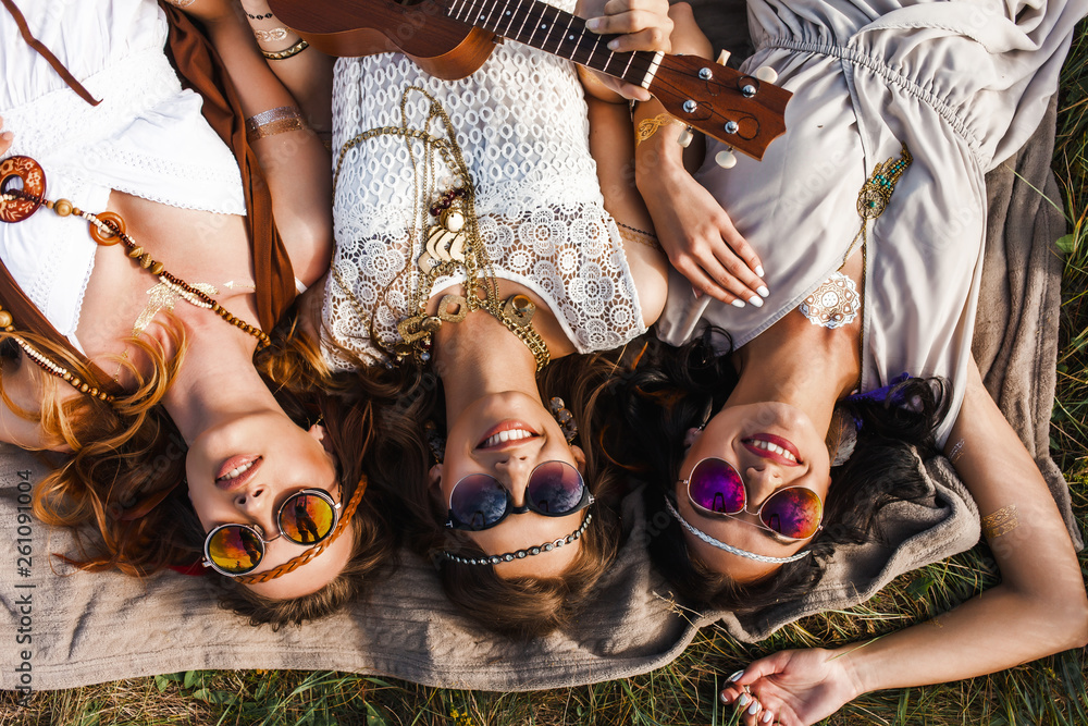 Obraz na płótnie Three cute hippie girl lying on the plaid outdoors, best friends having fun and laughing, play ukulele, sunglasses, feathers in their hair, bracelets, flash tattoo, indie, Bohemia, boho style top view w salonie