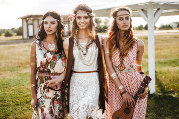 Three cute beautiful hippie girl in the setting sun, outdoors, the best of friends smiling and having fun, makeup, long hair, feathers in their hair, bracelets, flash tattoo, indie, Bohemia boho style