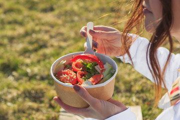 Smiling hipster woman having a relaxing lunch break outdoors, she is sitting on the grass and eating a salad bowl from paper box