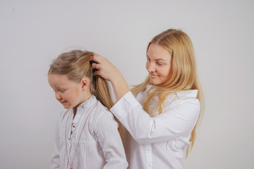 mom and daughter in white shirts with long blonde hair posing on a solid background in the Studio. a charming family takes care of each other and makes braids.