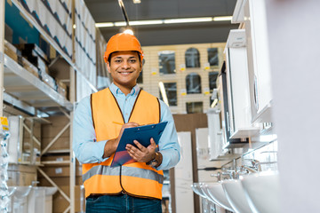 Wall Mural - cheerful indian warehouse worker writing in clipboard, smiling and looking at camera