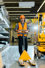 Wall Mural - cheerful warehouse worker standing with pallet jack, smiling and looking at camera