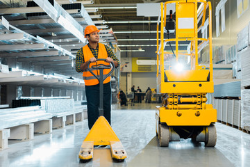 Wall Mural - serious indian worker in safety vest and helmet standing near pallet jack in warehouse