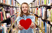 Valentine's Day, Love And Education Concept - Smiling Pretty Teenage Girl In Denim Jacket Holding Red Heart Over Library Background