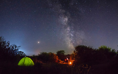 Night camping. Couple tourists have a rest at a campfire near illuminated tent under amazing night sky full of stars and milky way. Low light.