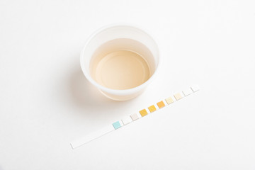 A combination of urine sample in a small round plastic container with a urine reagent strip deliberately and artistically set on a plain white background.