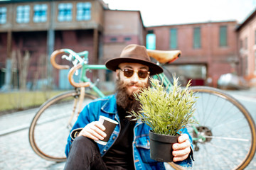 Wall Mural - Portrait of a stylish hipster with flower pot sitting near his retro bicycle on the street on the urban background