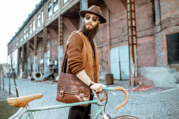 Wall Mural - Lifestyle portrait of a bearded hipster dressed stylishly walking with retro bicycle on the industrial urban background