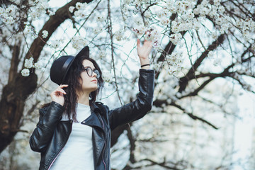 Outdoor spring portrait of fashion sensual young girl posing in amazing blooming garden. Wearing a hat and leather jacket, feminine look, elegant glamorous style. On the background of cherry blossoms