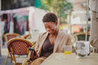 © Miguel Valls/ADDICTIVE STOCK - Cheerful African American elegant woman holding mobile phone and sitting at table with glass of juice near baggage in street cafe