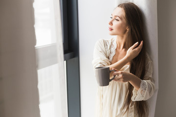 Canvas Print - Attractive young girl in night clothes standing by the window with a mug of coffee