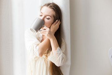 Canvas Print - Attractive young girl in nightwear stands at the window and drinks coffee. Morning mood