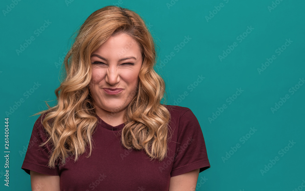 A young woman is squinting a sour grimace while standing against blue background. - obrazy, fototapety, plakaty 