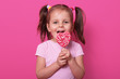 © sementsova321 - Happy cute girl wears rose t hirt, stands isolated over pink background, holds bright lollipop in hands. Cheery child with opened mouth tasting delicious candy. Childhood and tastes concept.
