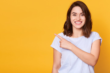 Close up portrait of nice cute adorable smiling charming cheerful girl pointing with her index finger aside, copy empty space for promotion text or advertisment, isolated over yellow background.