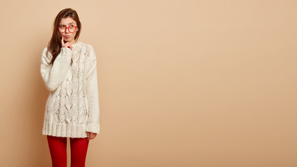 Wall Mural - Beautiful girl wonders if she can afford little rest, stands in thoughtful pose, folds llips and touches cheek, focused away, wears long white jumper and red tights, isolated over beige background.