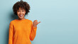 Leinwandbild Motiv Photo of delighted cheerful Afro American woman with crisp hair, points away, shows blank space, happy to advertise item on sale, wears orange jumper, demonstrates where clothes shop situated