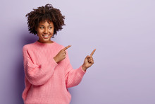 Studio Shot Of Attractive Young Female With Curly Hair, Toothy Smile, Healthy Dark Skin, Points Aside On Free Space, Demonstrates Something Awesome Against Purple Copy Space, Wears Pink Jumper