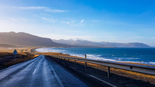An Endless Road Along The Coast. Road Stretches Over The Horizon. Waves Gently Wash The Shore. Empty Road, With No Cars Pasing By. In The Back Tall Mountains Emerge From The Seashore. Road Trip.