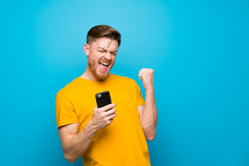 Redhead man over blue wall with phone in victory position