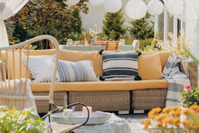 Real Photo Of A Yellow, Garden Sofa With A Table And Lamps In The Background. Close-up Of Plants