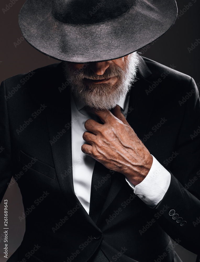 An old male actor playing an English spy role, dressed in black suit,  hiding behind fedora hat, having mysterious elegant look. Isolated studio  portrait over dark background Stock-foto | Adobe Stock