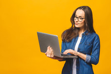 Young Happy Smiling Woman In Casual Clothes Holding Laptop And Sending Email To Her Best Friend. Isolated Against Yellow Background.