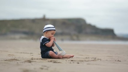 Sticker - Baby boy sitting on the beach near the water and plays with a toy ship and teddy bear