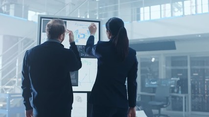 Wall Mural - Male and Female Design Engineers are Working and Discussing Something on a Interactive Whiteboard Next to an Electric Car Chassis Prototype. In High Tech Laboratory Facility with Vehicle Frame.