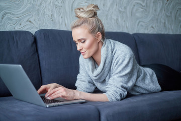 Happy blond woman lying prone on sofa and working on laptop computer