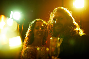 Wall Mural - Jolly beautiful young couple with long curly hair standing in lights and smiling at camera while spending time at party