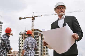 Building inspector. Portrait of experienced elderly engineer in formal wear and white helmet holding a project plan and checking work while standing at construction site.